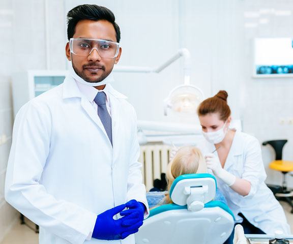 A male dentist wearing gloves, glasses, and a white coat standing and poses at his dental clinic.