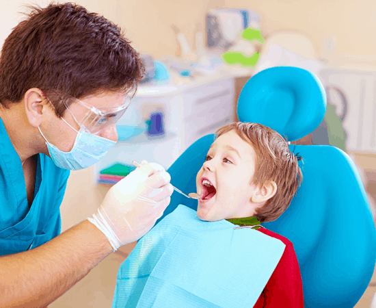 A male dentist with glasses examining the teeth of a young kid seated in the dental chair