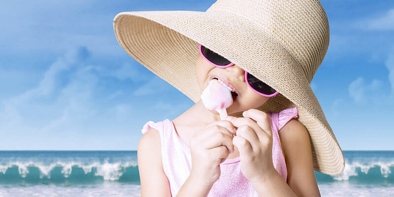 Little girl standing on the beach while wearing a swimsuit and hat, enjoying ice cream